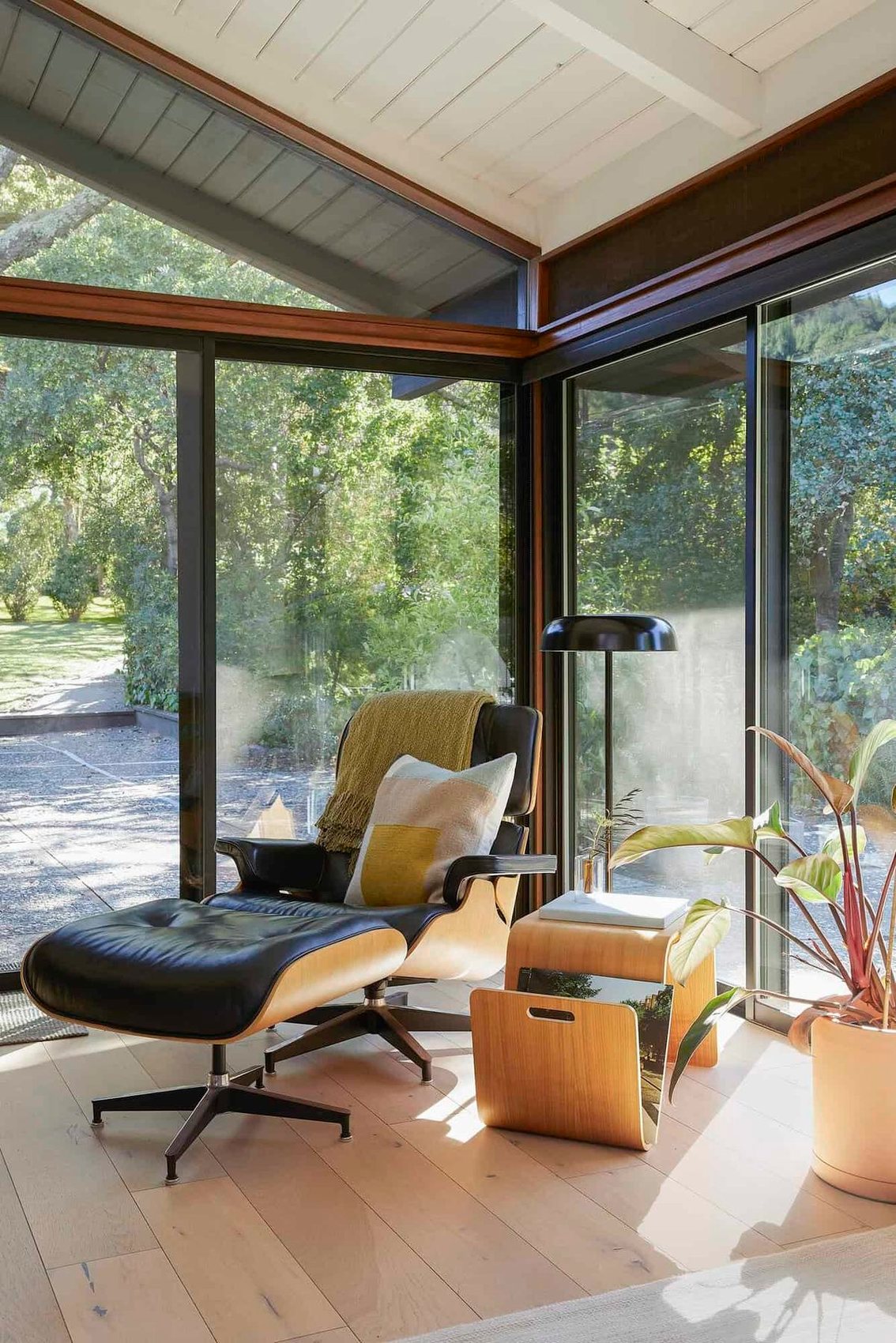 An Eames lounge chair by floor-to-ceiling windows looking into trees