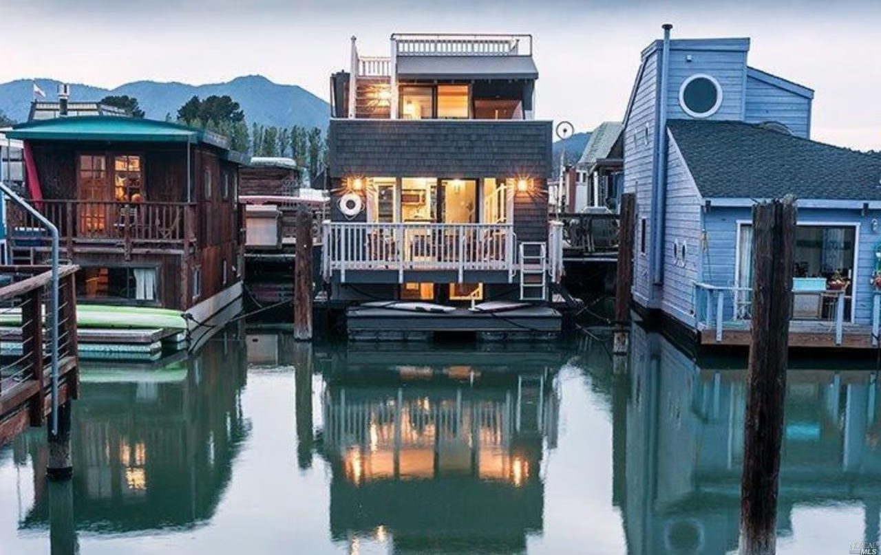 Sausalito houseboats at dusk, their lit windows reflected in still water below the hills