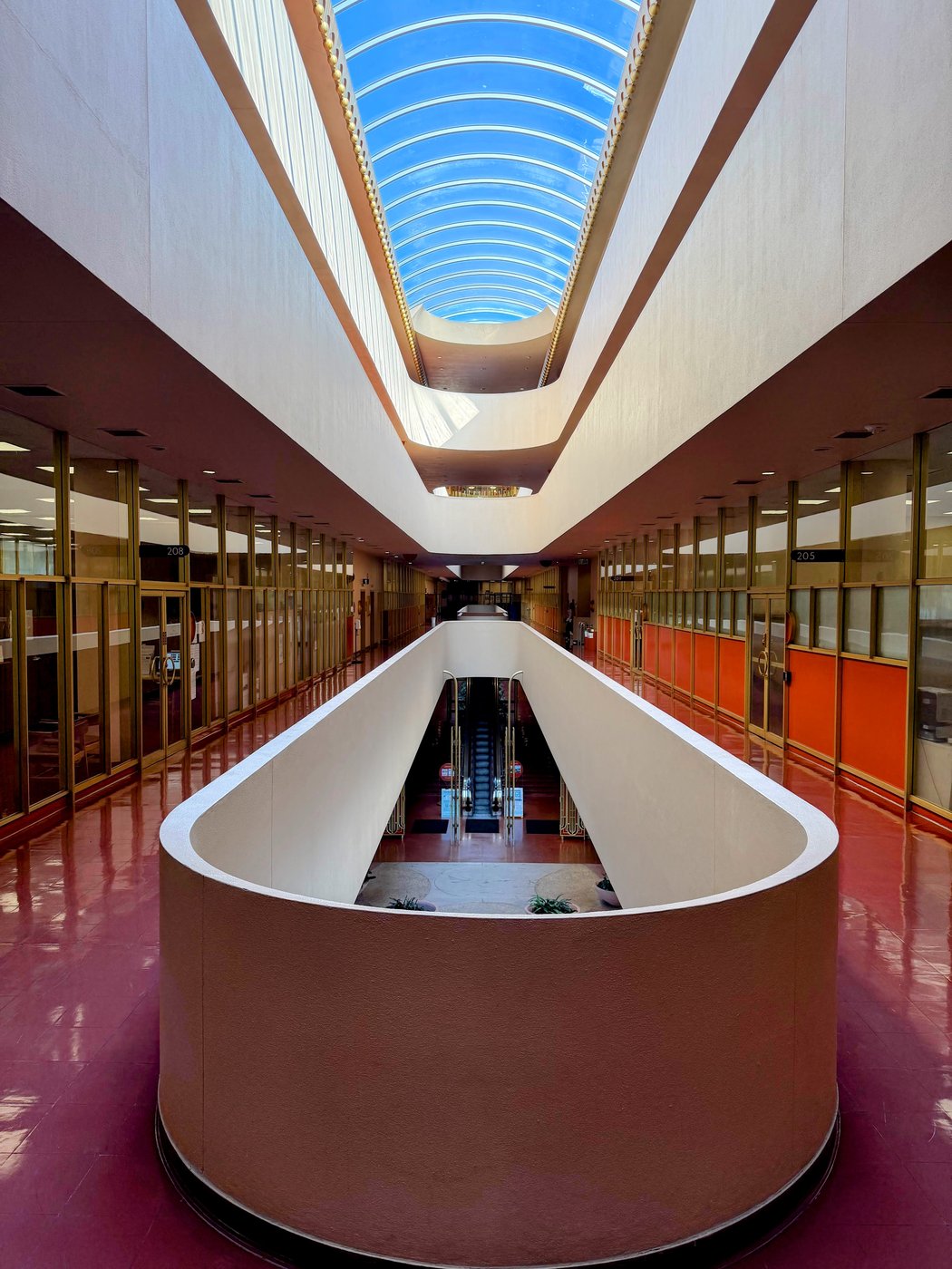 The skylit central corridor of the Marin County Civic Center, a long barrel vault of glazing overhead, gold screens and numbered office fronts down both sides, and a curved white balcony wall enclosing the escalator well, all over a polished terracotta floor