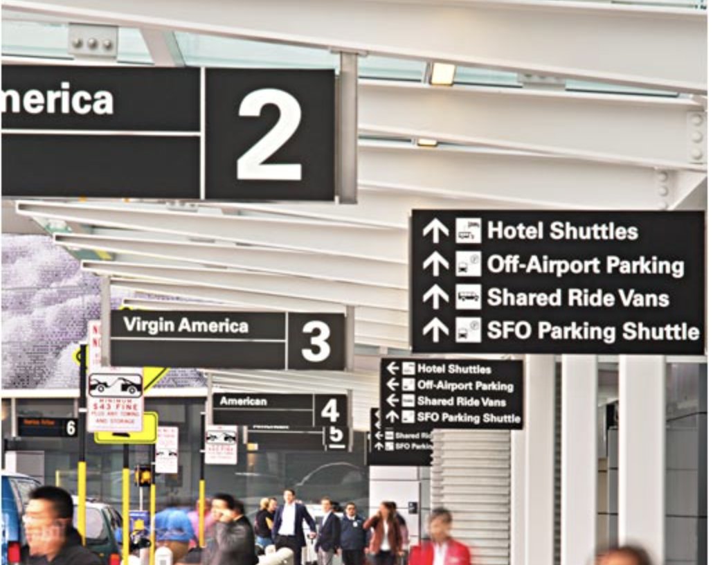 Overhead terminal signage at SFO, airline names over numbered doors beside a stacked white-on-black panel directing to hotel shuttles, off-airport parking, shared ride vans and the SFO parking shuttle