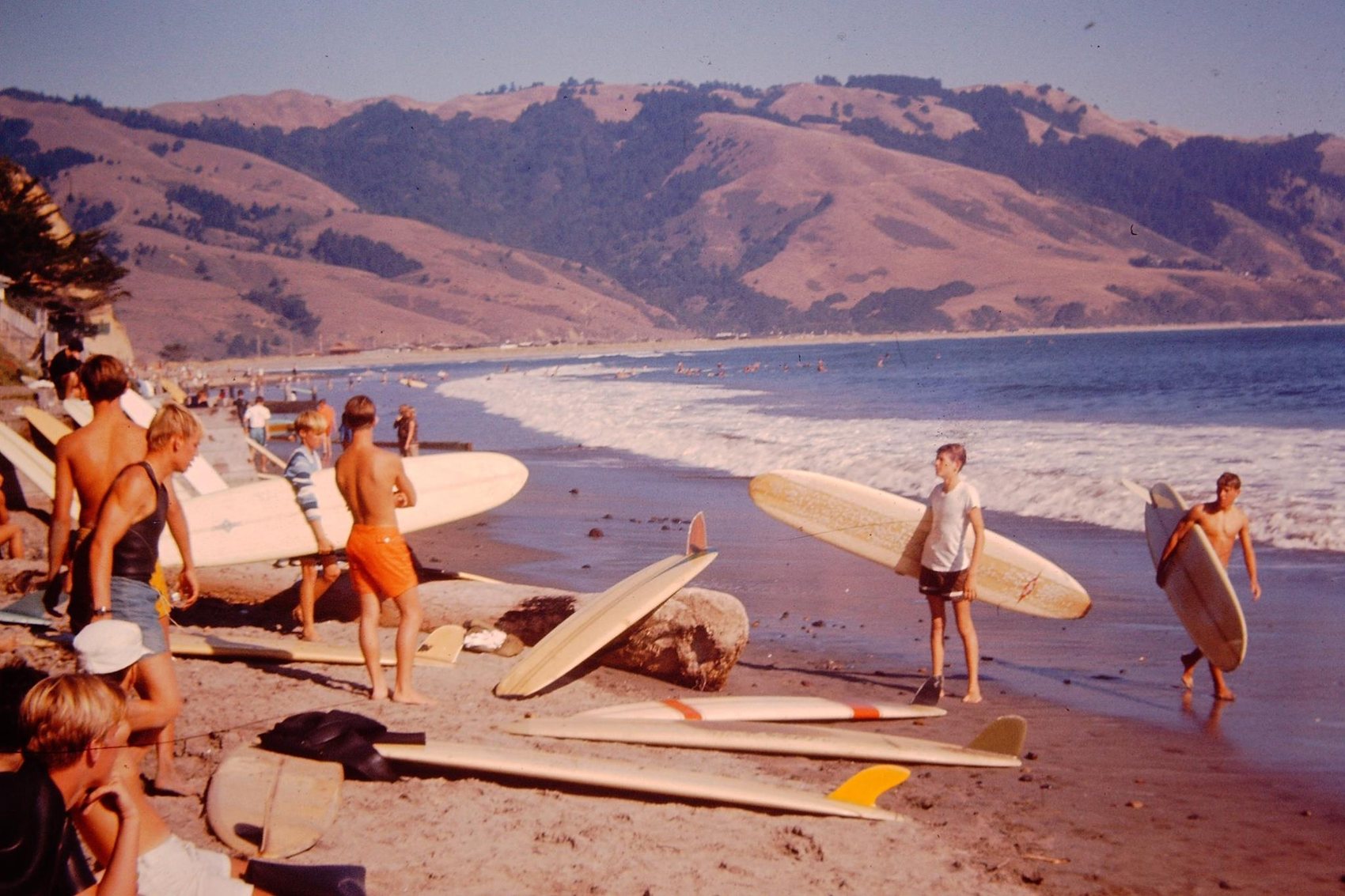 A 1966 beach scene of surfers and longboards on the sand at Bolinas, hills behind the bay